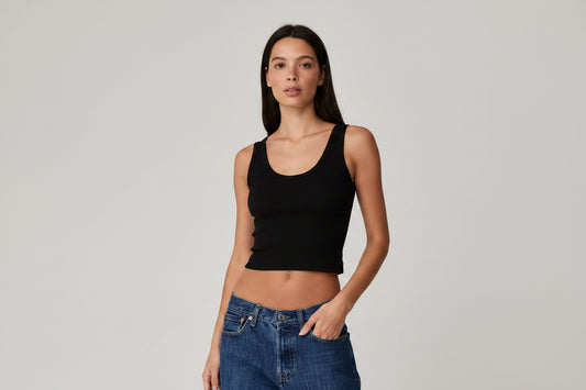 A woman modeling a fitted white scoop-neck crop top paired with blue jeans, photographed against a plain light gray studio background.
