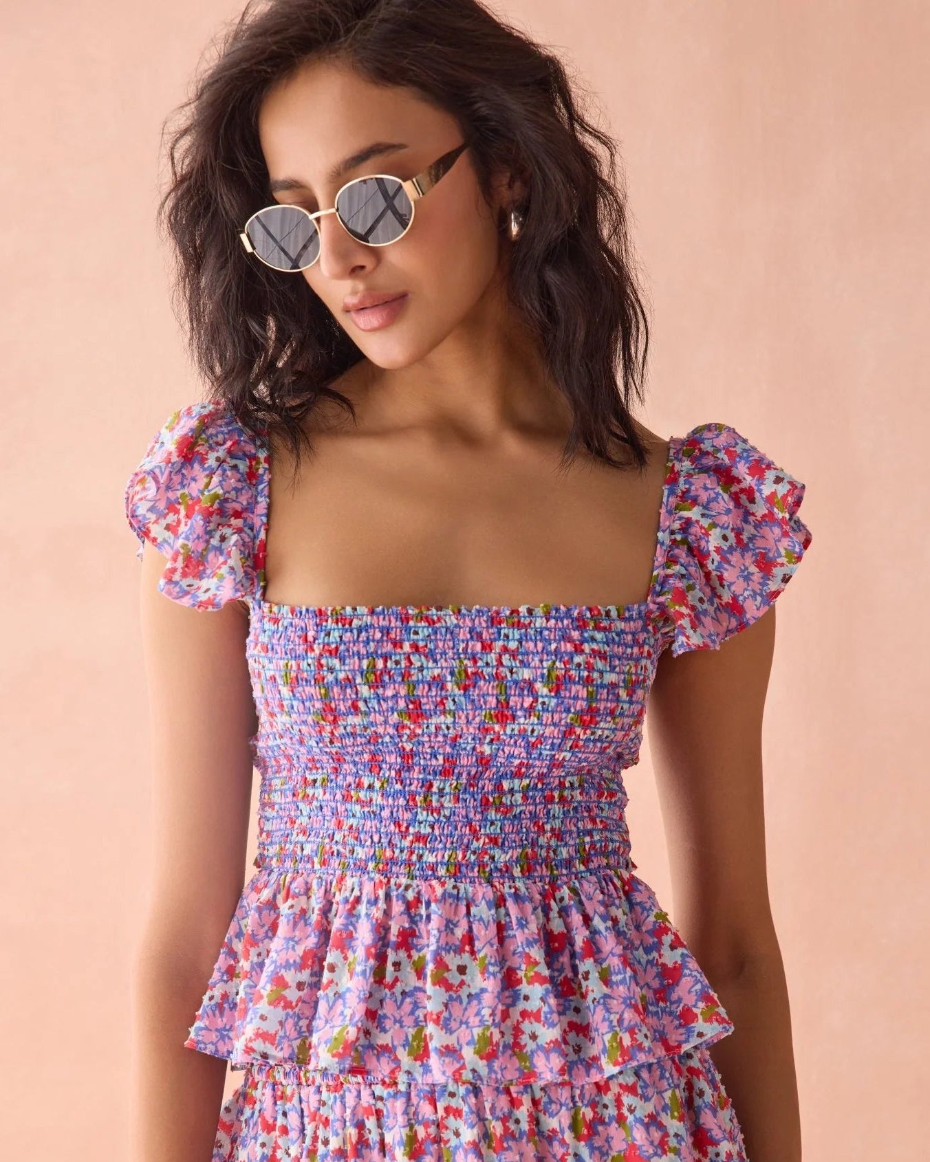A woman wearing a colorful floral smocked dress with puff sleeves and round sunglasses, posed against a solid peach-pink background.