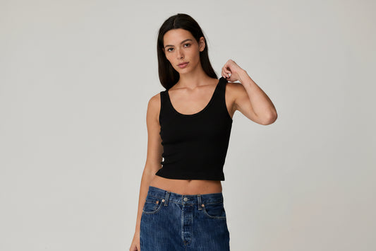 A woman modeling a fitted white ribbed tank top paired with dark blue jeans, photographed against a plain light gray studio background.