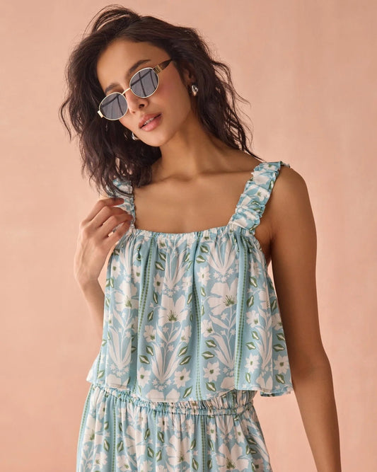 A woman wearing a light blue sleeveless dress with a white floral print and square neckline, posed against a solid peach-pink studio background.